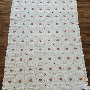 May include: A rectangular off-white crochet blanket featuring a repeating grid pattern of square fabric patches. Each patch is embroidered with a small red strawberry with green leaves. The blanket has a scalloped crochet border edge.