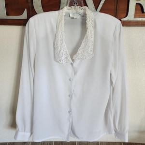 May include: A white long-sleeved blouse with a delicate lace collar. The blouse has a button-down front and is hanging on a clear hanger. The background features a wooden sign with the word "LOVE" in metal letters.