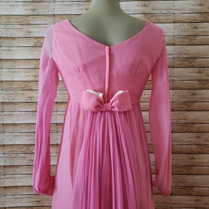 May include: A long-sleeved pink dress featuring a V-neckline and a central vertical zipper closure. The back of the dress is accented with a prominent pink bow at the waist, from which a sheer, pleated fabric panel cascades down the length of the skirt.