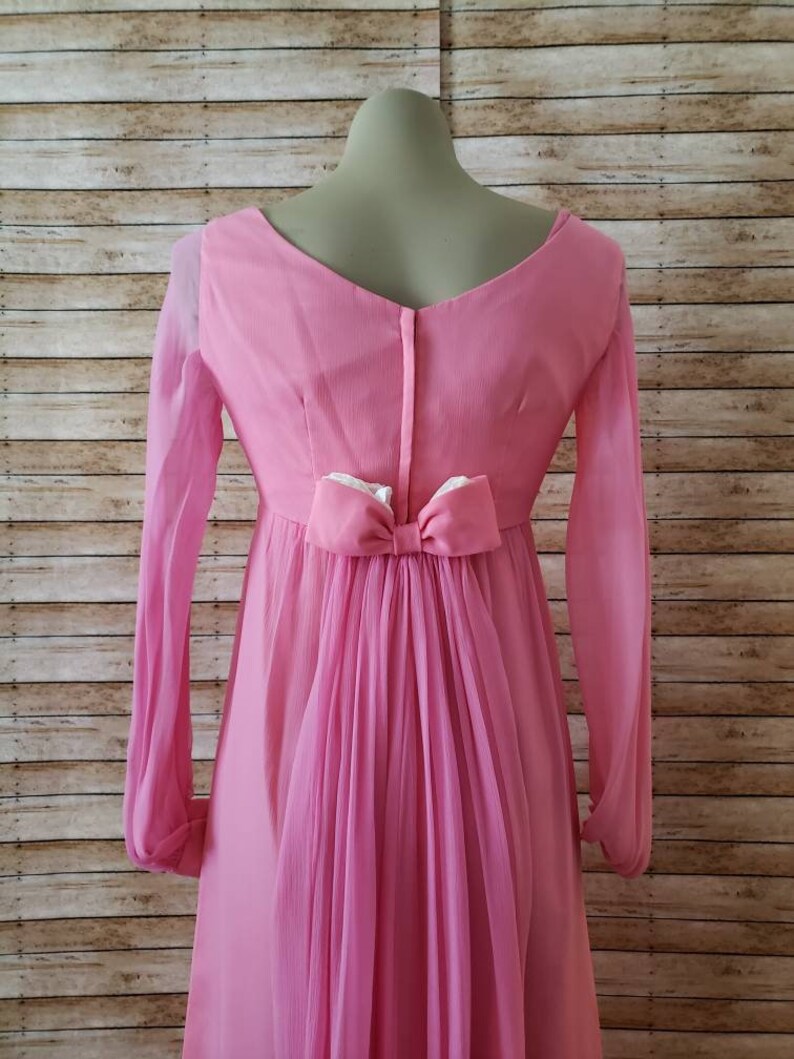 May include: A long-sleeved pink dress featuring a V-neckline and a central vertical zipper closure. The back of the dress is accented with a prominent pink bow at the waist, from which a sheer, pleated fabric panel cascades down the length of the skirt.