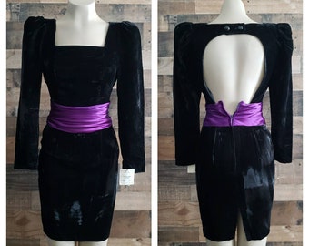May include: A black velvet dress with long sleeves and a square neckline. The dress features a vibrant purple satin sash at the waist. The back of the dress has a keyhole design. The dress is displayed on a mannequin.