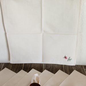 Pu&ograve; includere: Tovaglioli di lino bianco sporco, alcuni piegati e disposti con un anello portatovagliolo in legno. Un tovagliolo &egrave; appeso a una clip di metallo. Un piccolo ricamo floreale in rosa e verde &egrave; nell'angolo di un tovagliolo.