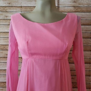 May include: A long-sleeved dress in a solid light pink color. The garment features a rounded neckline, a fitted bodice with vertical darts, and a flowing skirt. The fabric has a subtle, fine-textured, crinkled appearance throughout the entire piece.