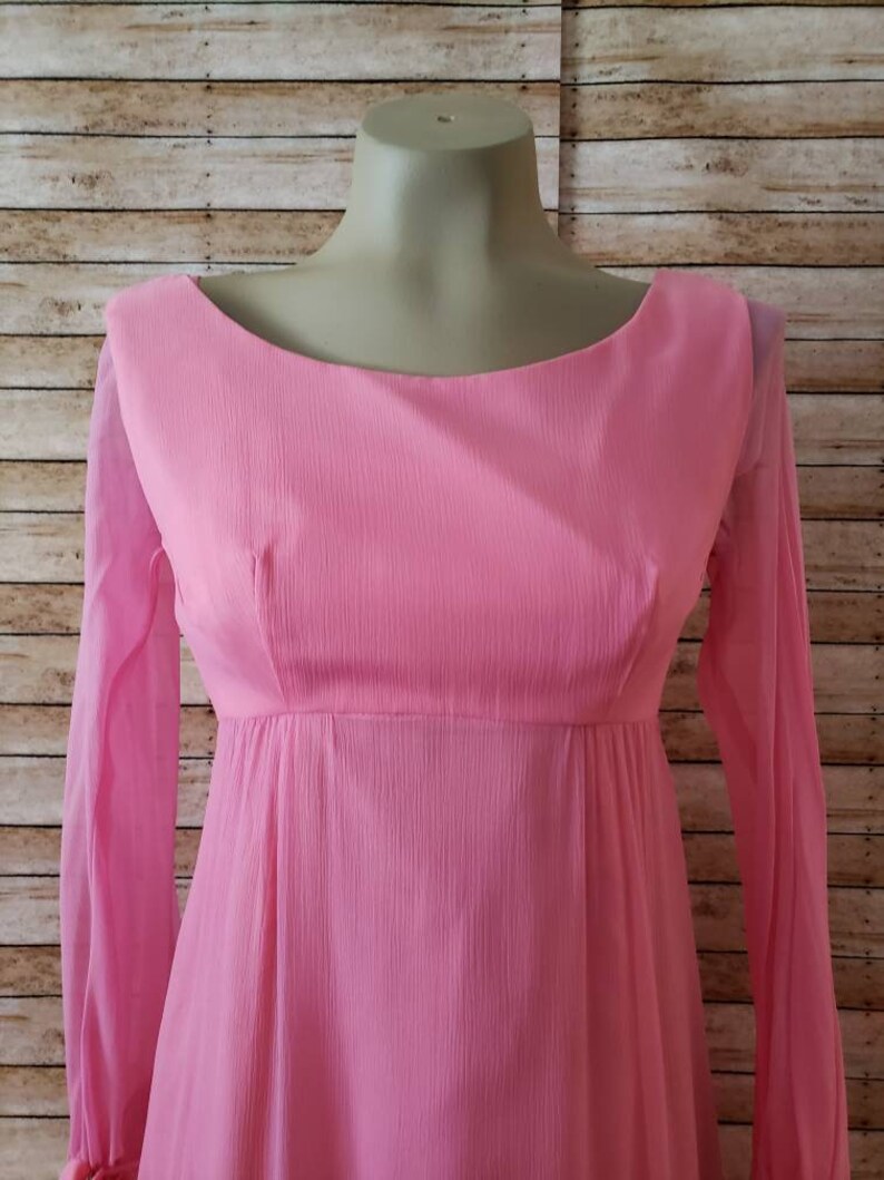 May include: A long-sleeved dress in a solid light pink color. The garment features a rounded neckline, a fitted bodice with vertical darts, and a flowing skirt. The fabric has a subtle, fine-textured, crinkled appearance throughout the entire piece.