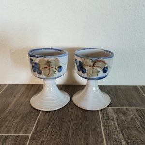 May include: Two white ceramic goblets with blue and green grapevine designs. The goblets have a white glaze and a slightly flared rim.