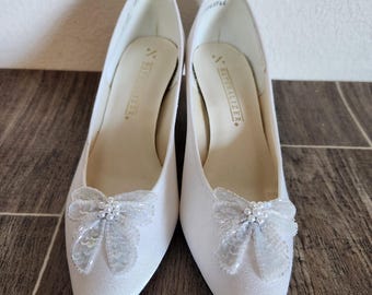 May include: A pair of white, pointed-toe pumps with a low heel. Each shoe features a decorative, translucent bow with bead accents on the toe. The shoes are set against a brown wooden surface.