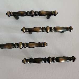 May include: Four antique bronze cabinet pulls. Each pull has a central, decorative design with rounded ends and a dark bronze finish. The pulls are arranged on a light background, showcasing their details and design.
