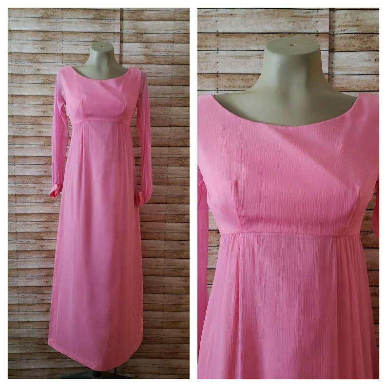 May include: A floor-length, solid pink dress featuring long, sheer sleeves with cuffs, a high empire waistline, and a wide, rounded scoop neckline. The fabric has a subtle, vertical crinkled texture throughout the bodice and the skirt.