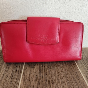 May include: A vibrant red leather wallet with a flap closure. The wallet features a decorative embossed design on the flap. The wallet is rectangular and appears to be closed. The wallet is on a wooden surface.