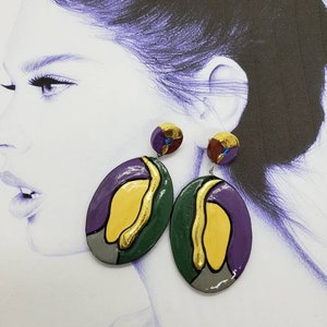 May include: A pair of oval-shaped earrings with a colorful abstract design. The earrings are made of green, purple, yellow, and gray clay with gold accents.