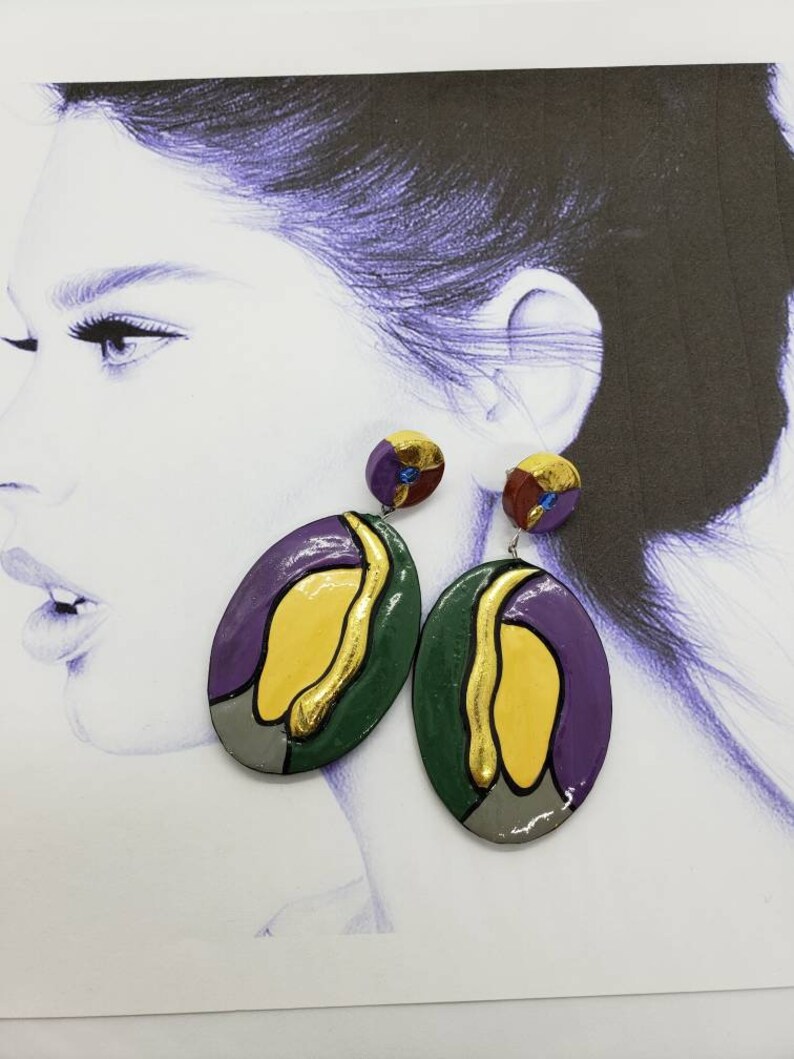 May include: A pair of oval-shaped earrings with a colorful abstract design. The earrings are made of green, purple, yellow, and gray clay with gold accents.