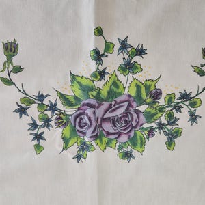 May include: Cream-colored fabric with a floral design. The design features two purple roses surrounded by green leaves and small blue flowers. The floral arrangement is centered on the fabric.