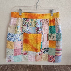 May include: A patchwork apron with a yellow, orange, and white floral print. The apron has a large pocket in the center and is hanging on a clear plastic hanger.
