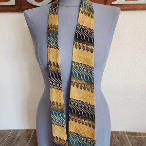May include: A patterned necktie displayed on a mannequin. The tie features a repeating design of blue, brown, and gold shapes. The tie is made of a silky material and is displayed against a neutral background.