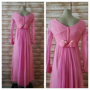 May include: A floor-length pink dress featuring long sheer sleeves, a V-neckline, and a central back zipper. The waist is accented with a decorative pink bow, from which a sheer, pleated fabric panel extends downward to create a flowing train effect.