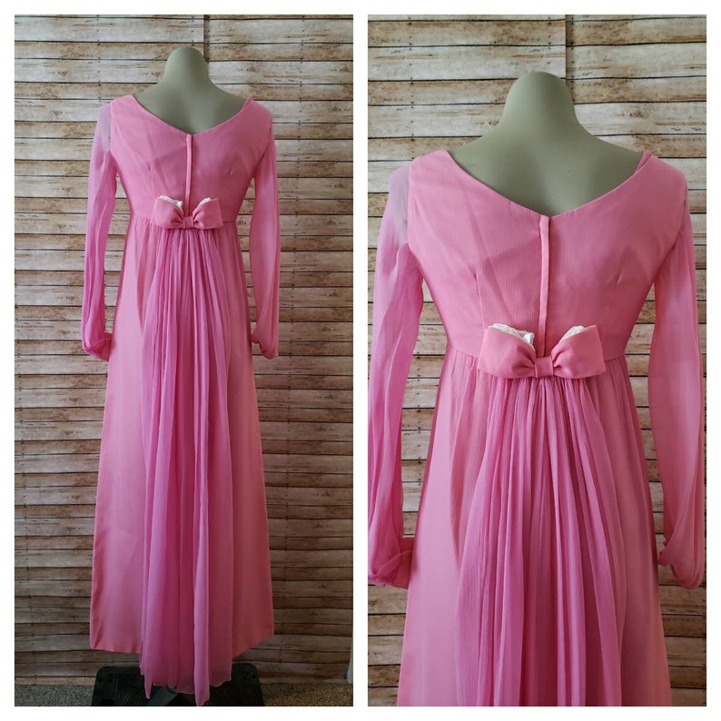 May include: A floor-length pink dress featuring long sheer sleeves, a V-neckline, and a central back zipper. The waist is accented with a decorative pink bow, from which a sheer, pleated fabric panel extends downward to create a flowing train effect.