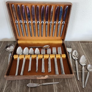 May include: A vintage silverware set in a wooden case. The set includes knives, forks, spoons, and serving utensils. The silverware has a textured handle design. The case has a brown exterior and a gold interior.