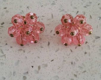 May include: A pair of pink flower-shaped earrings made of small, faceted glass beads. The beads are strung on a gold wire and have a gold-toned metal backing.