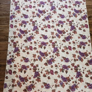 May include: A white tablecloth with a repeating pattern of purple grapes and brown leaves. The tablecloth has a white border.
