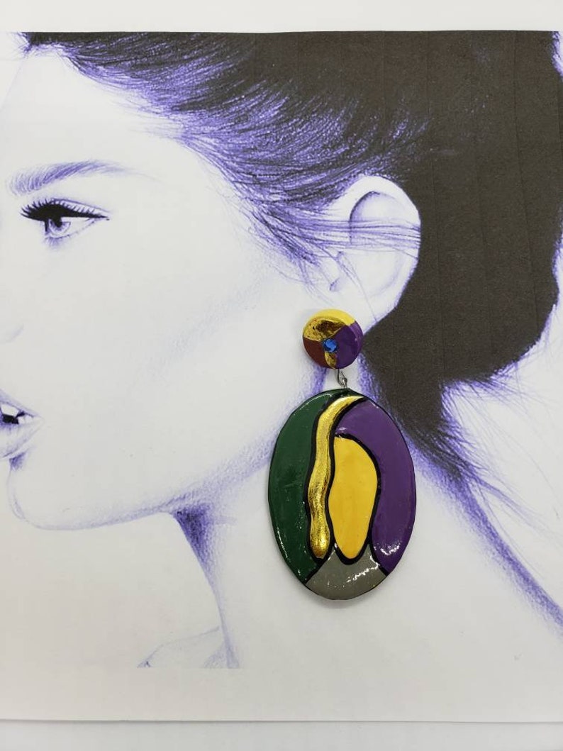 May include: A pair of colorful, abstract oval-shaped earrings. The earrings are made of clay and feature a green, yellow, purple, and gray color scheme. The earrings are hanging from a silver hook.