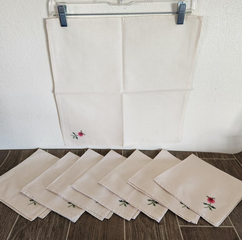Pu&ograve; includere: Un set di otto tovaglioli di lino bianco sporco, ciascuno con una piccola rosa rosa ricamata e foglie verdi. Un tovagliolo &egrave; appeso a un appendiabiti di plastica trasparente. I tovaglioli sono piegati e disposti su una superficie di legno.
