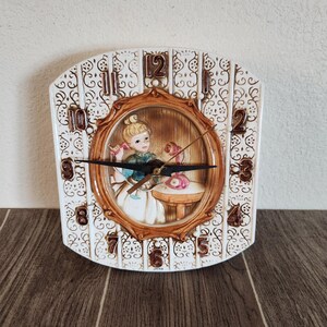 May include: A ceramic wall clock with a cream-coloured, embossed floral pattern and brown numerals. The centre features a three-dimensional scene of a person with blonde hair talking on a pink telephone. The clock has black hands and says JAPAN.