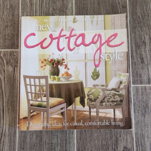 May include: A book titled "New Cottage Style" by Better Homes and Gardens. The cover features a photo of a living room with a green armchair, a white chair, a small table with a green tablecloth, and a vase of flowers. The text "Decorating ideas for casual, comfortable living" is at the bottom of the cover.