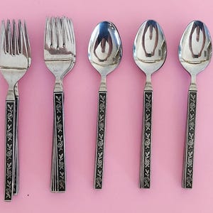 May include: Silverware set featuring two forks and three spoons. The utensils have black handles with a floral design. The background is a solid pink color.