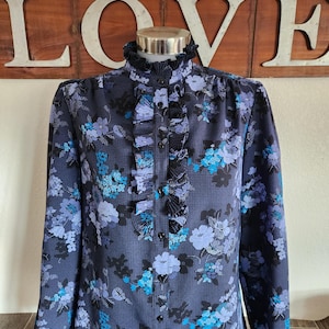 May include: A navy blue blouse with a floral pattern in shades of blue and black. The blouse features a high ruffled collar, a button-down front, and long sleeves. The floral print is composed of various flower shapes.