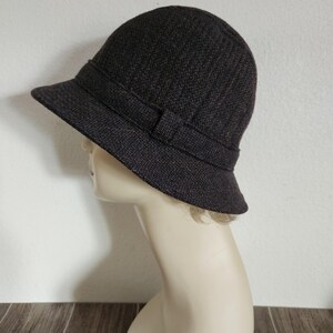 May include: A dark brown cloche hat with a textured weave. The hat features a band with a small bow detail. The brim is slightly angled, and the hat is displayed on a mannequin head.