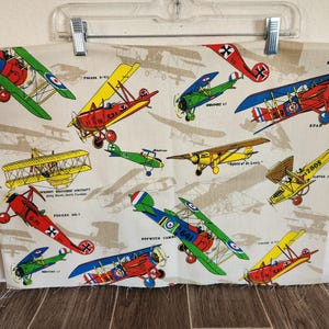 May include: Cream-colored fabric featuring a repeating pattern of vintage biplanes in various colors, including red, green, yellow, and blue. The planes have propellers and are set against a backdrop of faint airplane silhouettes. Some planes have text labels.