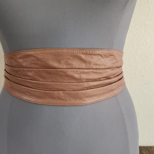 May include: A wide, light brown leather belt is wrapped around a mannequin. The belt has a textured appearance and is designed to be worn around the waist. The belt is a neutral color and appears to be made of genuine leather.