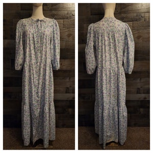 May include: A long, white dress with a floral print in shades of purple and green. The dress has a button-up front, a gathered waist, and a tiered skirt.