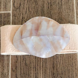 May include: A peach-colored elastic belt with a large, oval, iridescent shell-like buckle. The buckle has a pearlescent sheen with pink and white tones. The belt is set against a brown and tan wood-grain background.
