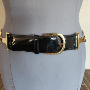 May include: Black patent leather belt with gold-tone metal hardware. The belt features a gold buckle and decorative gold accents on each side. The belt is displayed on a mannequin.