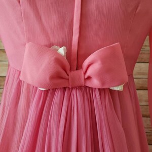 May include: A bright pink dress featuring a central vertical seam and a large, horizontal pink bow attached at the waist. The fabric has a fine, vertical pleated texture, and the bow reveals a small portion of white lining underneath its folds.