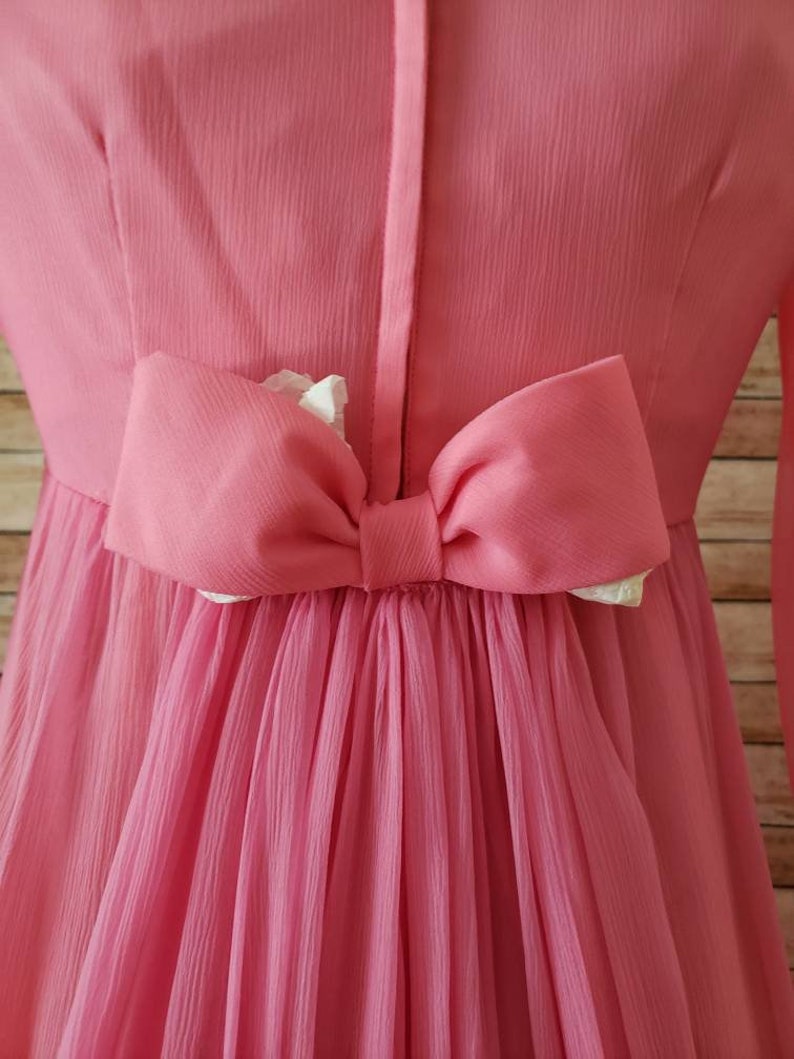 May include: A bright pink dress featuring a central vertical seam and a large, horizontal pink bow attached at the waist. The fabric has a fine, vertical pleated texture, and the bow reveals a small portion of white lining underneath its folds.