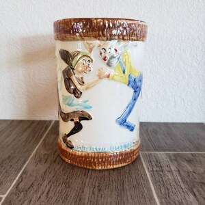 May include: A ceramic mug with a brown rim and base. The mug features a raised design of two people in a playful scene. The text "Mit Rug Cutting" is visible on the base.