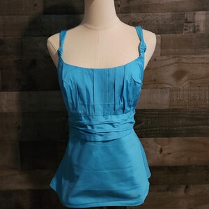 May include: A bright turquoise sleeveless top featuring a square neckline, vertical pleats across the bodice, and a gathered waist detail. The garment includes wide shoulder straps with knotted accents at the top and a flared hemline.