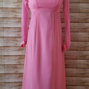 May include: A floor-length, solid pink dress featuring long, sheer sleeves with cuffed wrists, a rounded neckline, and an empire waistline. The garment is displayed on a mannequin and has a flowing, straight-cut skirt that reaches the floor.