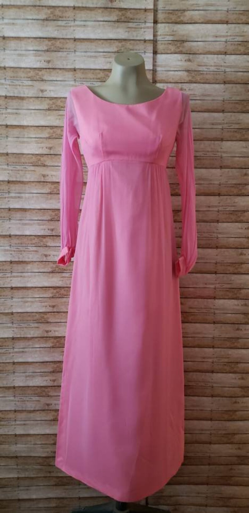 May include: A floor-length, solid pink dress featuring long, sheer sleeves with cuffed wrists, a rounded neckline, and an empire waistline. The garment is displayed on a mannequin and has a flowing, straight-cut skirt that reaches the floor.