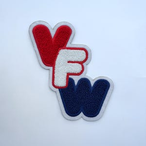 May include: A patch with the letters "VFW" in red, white, and blue. The letters are embroidered with a textured, fuzzy appearance and outlined in white. The patch is likely for a military or veterans organization.