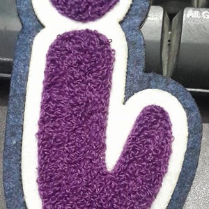 May include: A purple, embroidered letter "i" patch. The letter is filled with a textured, fuzzy material and outlined in white and navy blue. The patch is likely for clothing or accessories.