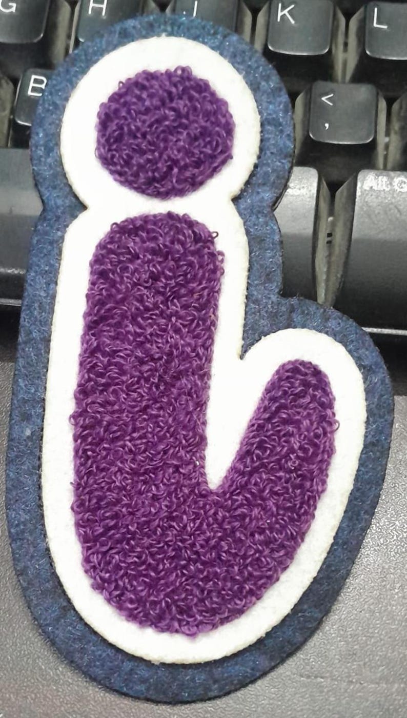 May include: A purple, embroidered letter "i" patch. The letter is filled with a textured, fuzzy material and outlined in white and navy blue. The patch is likely for clothing or accessories.