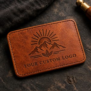 Può includere: Una toppa rettangolare in pelle marrone con angoli arrotondati caratterizzata da un design in rilievo raffigurante un sole che sorge sopra cime montuose e tre foglie alla base. Il testo YOUR CUSTOM LOGO è impresso in basso sulla toppa.
