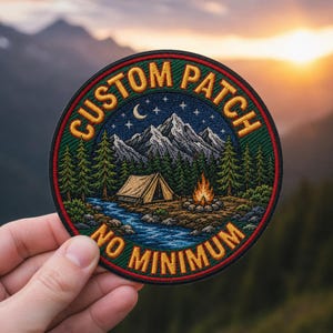 Pode incluir: Um emblema bordado circular apresentando uma paisagem de montanha à noite com uma tenda, fogueira e rio. O texto CUSTOM PATCH está arqueado no topo e NO MINIMUM na parte inferior em fio amarelo sobre uma borda verde escura.