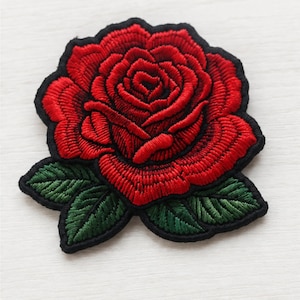 May include: A fabric patch featuring a detailed embroidered red rose with layered petals and two dark green leaves at the base. The entire design is outlined with a thick black border, creating a clear contrast against the surrounding area.