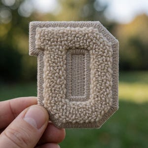 May include: A beige letter D patch featuring a textured, looped pile surface surrounded by a smooth, flat-stitched border. The letter is constructed with a dense, raised fabric texture that creates a distinct contrast against the flat, embroidered edge.