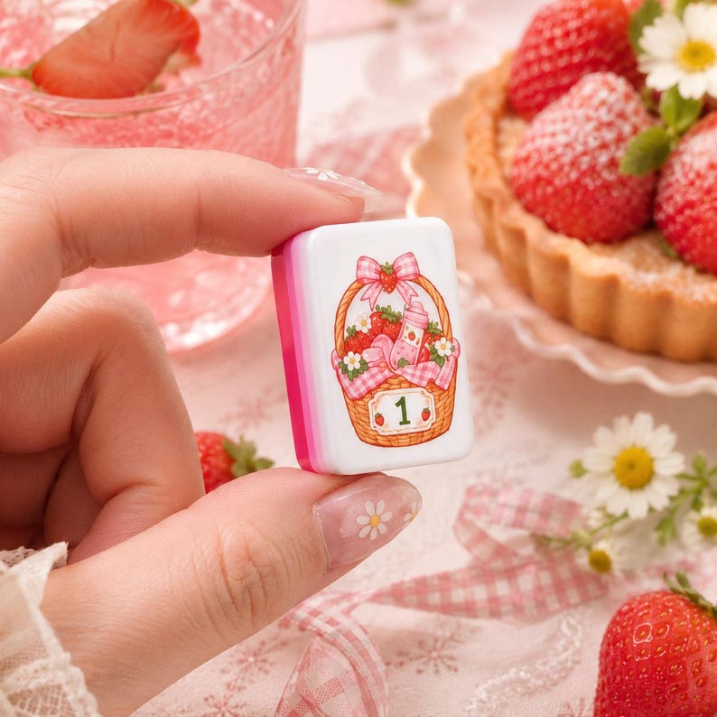May include: A rectangular white mahjong tile featuring a pink and brown illustration of a woven basket filled with strawberries, a small bottle, and white flowers, accented with a pink gingham bow and the green number 1 on the front surface.