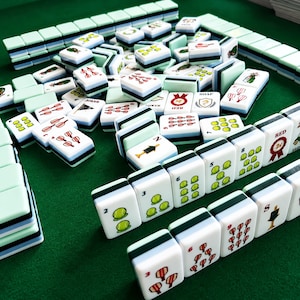 Pode incluir: Um conjunto de pe&ccedil;as retangulares de mahjong com faces brancas e lados em camadas nas cores verde-claro, azul e preto. As pe&ccedil;as apresentam v&aacute;rias ilustra&ccedil;&otilde;es coloridas, incluindo bolas de t&eacute;nis, fitas vermelhas e texto como RED, SOAP e JOKER numa superf&iacute;cie verde.